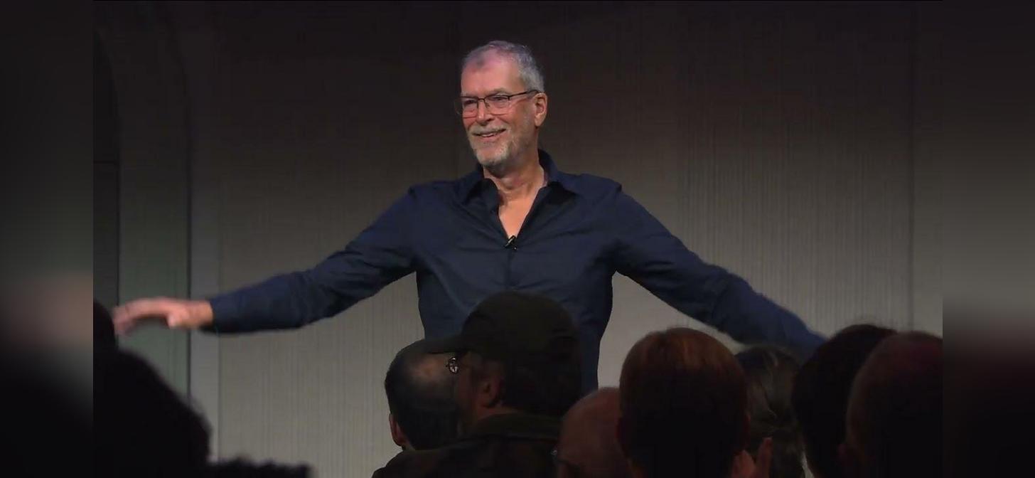 Man in blue shirt speaking on stage with arms outstretched, engaging audience.