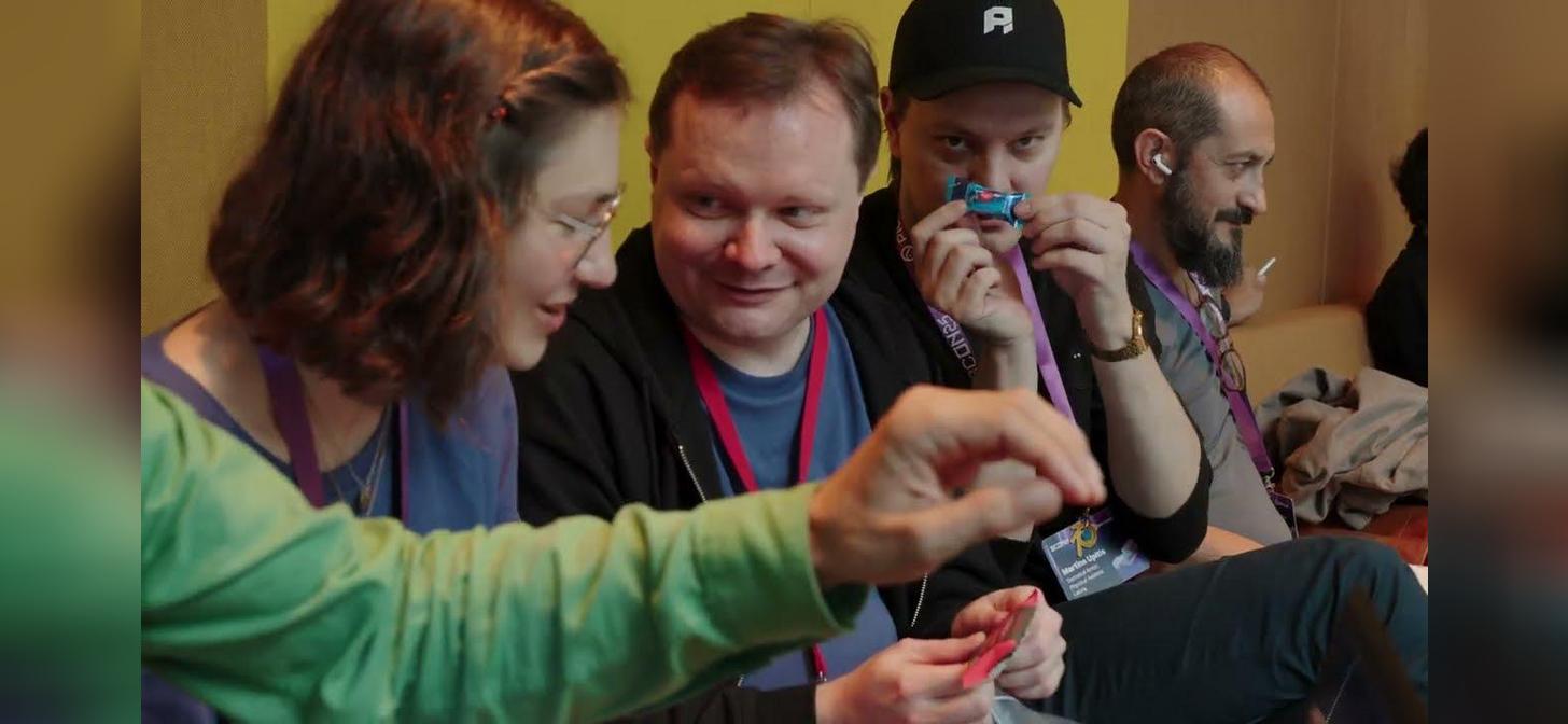 Four people wearing lanyards and badges are gathered, intently focused on a small object being held by the person on the left. The setting appears to be an indoor event or conference.